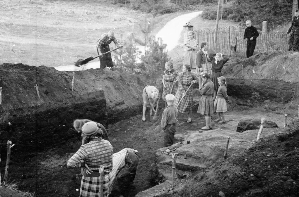 A black and white photograph of an excavation with adults and children taking part.