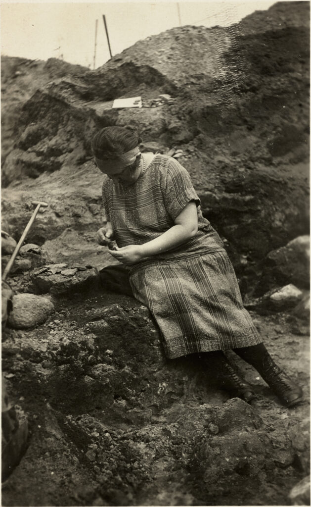 Sepia photograph of a woman in a excavation hole looking at finds.